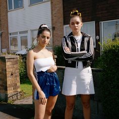 two young women standing next to each other in front of a house with their arms crossed