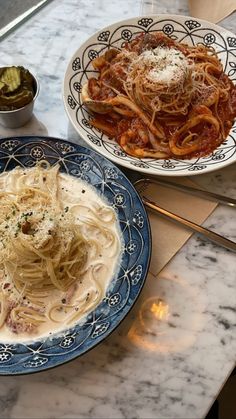 two plates of spaghetti and sauce on a marble counter top with silverware next to it