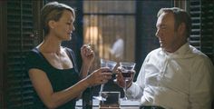 a man and woman sitting at a table with wine glasses in their hands, talking to each other