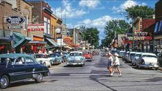 a woman walking down the middle of a street in an old fashioned town with cars parked on both sides