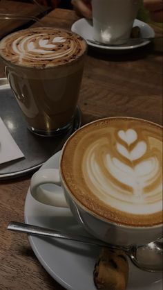 two cups of coffee on a table with spoons next to each other and one has a heart shaped latte in the foam