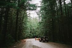 two people standing on the side of a dirt road in front of some tall trees