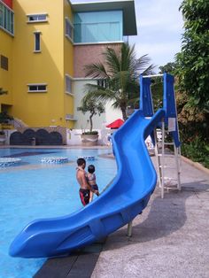 two children playing in a pool with a blue slide next to the swimming pool and yellow building