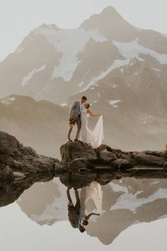 a bride and groom standing on a log in front of a mountain lake with their reflection in the water