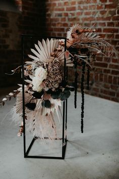 an arrangement of flowers and feathers in a vase on a table next to a brick wall