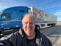 a man standing in front of a semi truck on the side of the road with a mustache