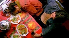 two men sitting at a red table with plates of food on it and one man leaning over the table