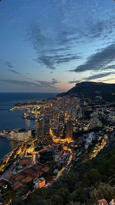 an aerial view of a city by the ocean at night with lights on buildings and mountains in the background