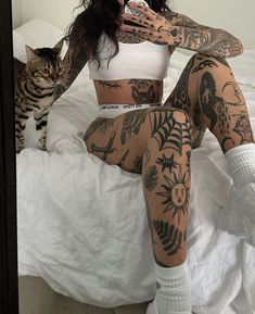 a woman with tattoos sitting on a bed next to a cat and holding a cell phone