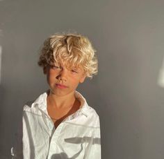 a young boy with blonde hair wearing a white shirt and standing in front of a gray wall