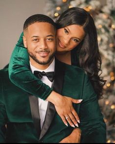 a man in a green suit and woman in a black dress hugging each other near a christmas tree