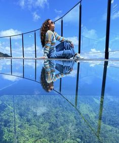 a woman sitting on the edge of a glass walkway over water with trees in the background