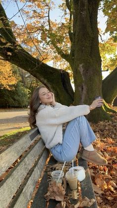 a woman sitting on top of a wooden bench next to a leaf covered tree and holding a cup