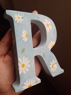 a person holding up a wooden letter with daisies on the letters and flowers painted on it