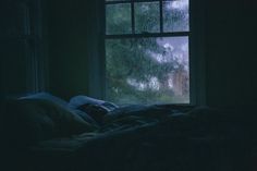 a bed in front of a window with rain coming down on the ground and trees outside