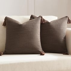 two brown pillows sitting on top of a white couch next to each other with tassels