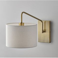 a wall light with a white shade on the side and a gold metal arm that is attached to it