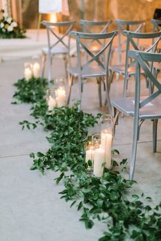 candles are lined up on the floor next to chairs with greenery growing down them