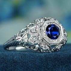 The intricate filigree on the band adds a unique and enchanting element, bringing a whimsical charm to the overall design of this vintage-inspired dome ring. Diamonds are skillfully integrated and evenly spaced to enhance the ring's visual appeal, creating a delightful balance and symmetry against the white gold backdrop. At the center of the ring, a captivating blue sapphire gemstone takes its place, expertly cut in a round brilliant style to maximize its brilliance and sparkle. CHARACTERISTICS Status: Made to order Origin: Thailand Metal: Solid 9K White Gold Ring Size: US 3-8 Total Gemstones Weight: 0.77 carat. (approx.) Total Gram Weight: 2.95 g. (approx.) *Pictures have been enlarged to show details* PRIMARY STONE(S) Stone: Natural Blue Sapphire Color: Blue Shape: Round Size: 5 mm. Num Fine Jewelry Sapphire Ring With Filigree, Elegant Blue Gemstone Dome Ring, Round Sapphire Filigree Jewelry, Anniversary Sapphire Ring With Filigree Fine Jewelry Style, Anniversary Fine Jewelry Sapphire Ring With Filigree, Elegant Domed Blue Sapphire Ring, Classic Blue Domed Jewelry, Elegant Blue Domed Sapphire Ring, Fine Jewelry Sapphire Ring With Filigree Design