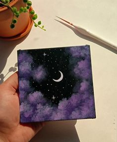 a hand holding up a small square piece of paper with the moon and stars painted on it