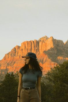 a woman standing in front of a mountain with trees and grass on the ground next to her