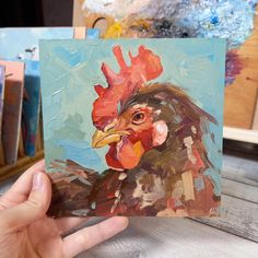 a painting of a rooster is being held up by someone's hand in front of some paintbrushes