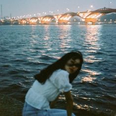 a woman sitting on the ground next to water with a bridge in the background at night