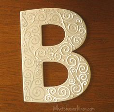 the letter b is decorated with swirly white paint on a wooden surface, and sits next to a wood table