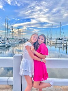 two girls hugging each other while standing on a pier with boats in the water behind them