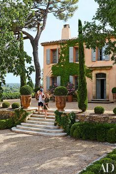 two people standing in front of a large house with lots of greenery on it