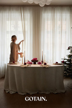 A Christmas-themed dining room with a festive table setting, a glowing Christmas tree, and sheer linen curtains in sand filtering soft natural light.