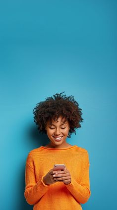 a woman smiling and looking at her cell phone while standing against a blue wall with her eyes closed