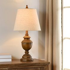 a lamp on a table next to a window with curtains and a book in front of it