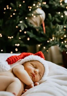 a baby wearing a santa hat sleeping in front of a christmas tree with lights on it