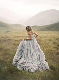 a woman in a dress is standing in a field with mountains behind her and grass on the ground