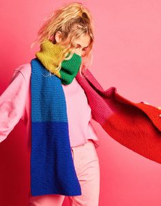 a woman with blonde hair wearing a multicolored scarf and pink sweater, standing against a pink background