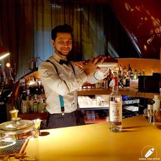 a man in a white shirt and bow tie standing behind a bar with bottles on it