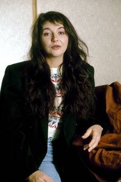 a woman sitting on top of a brown couch next to a white wall and wearing a black blazer