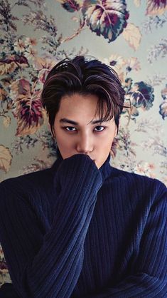 a young man covers his face with his hands while standing in front of a floral wallpaper