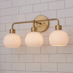 three light bathroom fixture with white glass globes on the back and brass finish, against a white brick wall