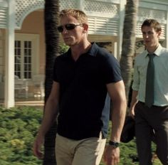 two men walking down the street in front of a building with palm trees and bushes