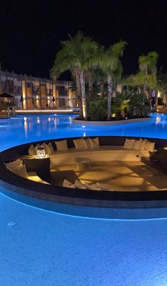 an empty swimming pool at night with lounge chairs and lights on the poolside, surrounded by palm trees