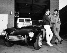 black and white photograph of two men standing next to an old sports car in front of a brick building