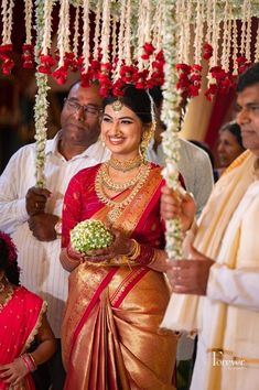 a woman in a red and gold sari holding a bouquet of flowers while standing next to other people