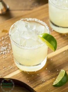 two margaritas sitting on top of a wooden cutting board next to lime wedges