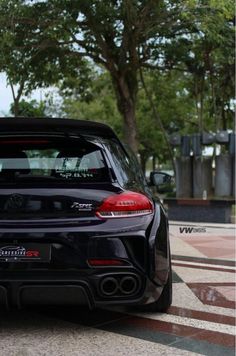 the back end of a black car parked in a parking lot next to some trees