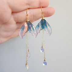 Discover the beauty of iridescent mermaid tail earrings in this stunning collection. These unique and eye-catching accessories are sure to make a splash wherever you go. Perfect for adding a touch of whimsy and magic to any outfit. Whether you're attending a wedding, a formal event, or simply want to add a touch of fierceness to your everyday look, these earrings are a versatile and stylish choice. 🍊Material: The earrings are made with shimmering resin mermaid tails, Czech glass water drops and the 14K gold plated (nickel-free) hooks. Handmade in Maryland, USA! 🌺Size: approx.(W x L) 2.54cm x 9.5cm 🎀Handmade jewelry: Each item is made to order, which gives our pieces a unique meaning that is specific and special to you.Therefore, there are no two items exactly alike. Contains small parts Diy Mermaid Accessories, Mermaid Jewellery, Magical Jewelry Fantasy Earrings, Iridescent Fantasy Jewelry For Parties, Fantasy Iridescent Jewelry For Parties, Iridescent Mermaid, Mermaid Jewelry Aesthetic, Mermaid Tail Aesthetic, Handmade Unique Iridescent Earrings