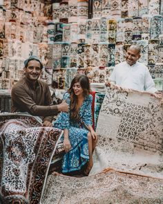 two men and a woman are sitting in front of rugs on the ground at an outdoor market