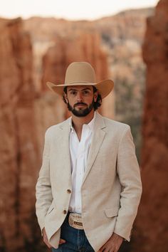 a man wearing a cowboy hat standing in front of some mountains and rocks with his hands in his pockets