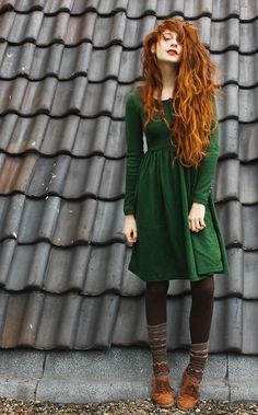 a woman with long red hair standing in front of a roof wearing green dress and brown boots